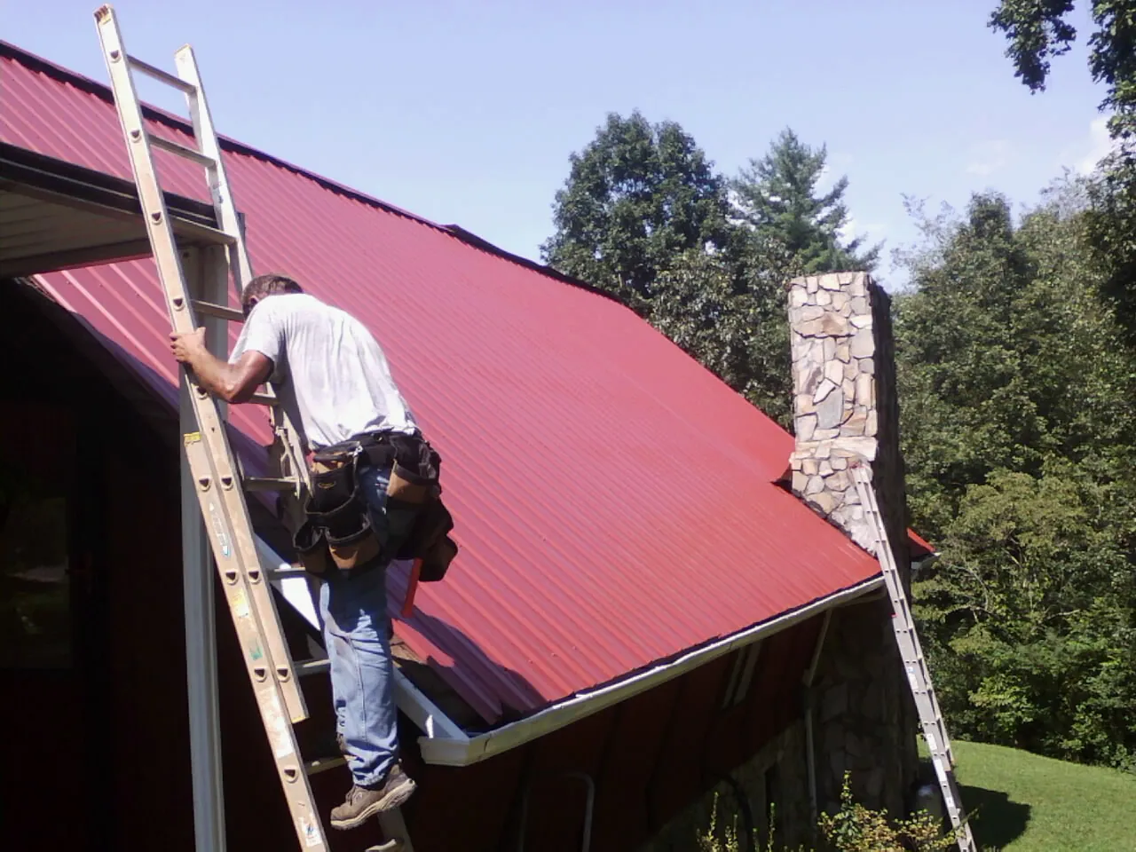 Roofer on a ladder beside a red metal roof and stone chimney — Soffit & Fascia Repair by Roofing Brothers in Niagara Falls
